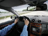 A person driving a car on a highway, holding the steering wheel with both hands. The view is from the back seat, showing the dashboard, road ahead, and side rails through the windshield.