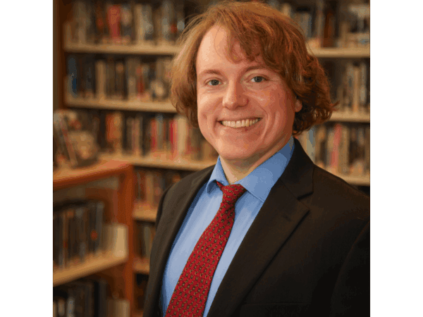 A smiling person with wavy light brown hair, wearing a black suit, blue shirt, and red tie, stands in front of bookshelves filled with books in a library.